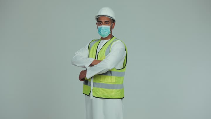 Reconstruction is done by the hands of the sons of the nation, wearing a mask for protection and prevention from viruses, an Arab Gulf Emirati man wearing a kandura, helmet and safety suit, standing with his hands crossed and looking at the camera with gestures of self-confidence, formal dress for work in the engineering sector and projects, white background