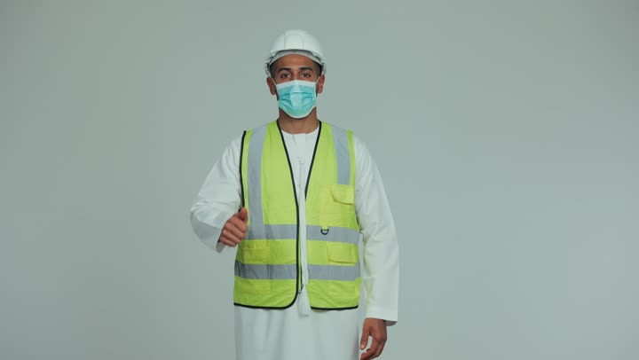 Wearing a mask for protection and prevention from viruses, an Arab Gulf Emirati man wearing a kandura, helmet and safety suit, raising his thumb to express quality and excellence, formal dress for work in the engineering and projects sector, white background