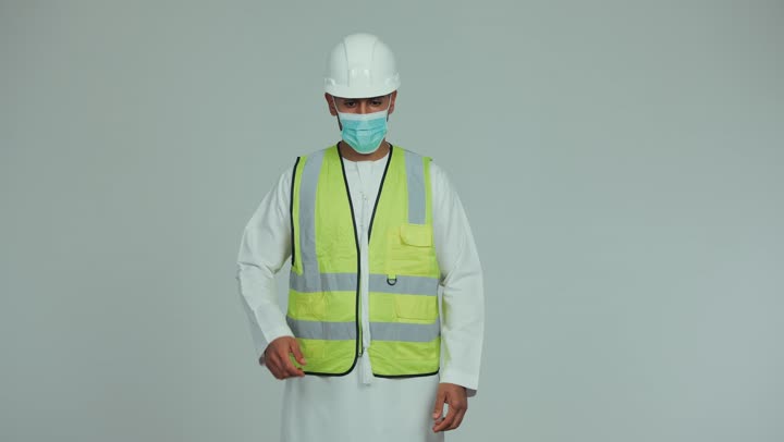 Reconstruction is done by the hands of the sons of the nation, wearing a mask for protection and prevention from viruses, an Arab Gulf Emirati man wearing a kandura, helmet and safety suit, standing with his hands crossed and looking at the camera with gestures of self-confidence, formal dress for work in the engineering sector and projects, white background