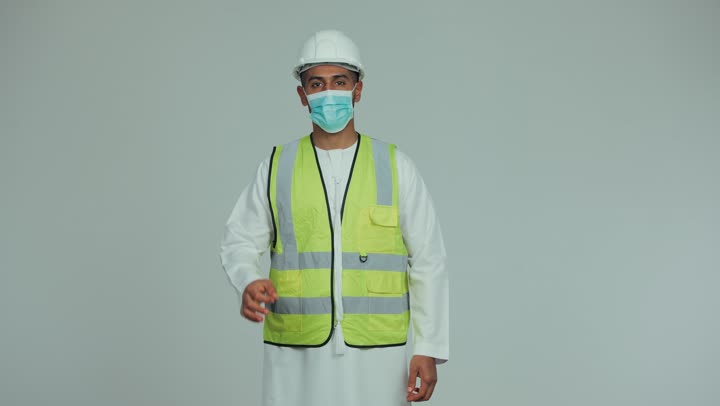 Wearing a mask for protection and prevention from viruses, an Arab Gulf Emirati man wearing a kandura, helmet and safety suit, raising his thumb to express quality and excellence, formal dress for work in the engineering and projects sector, white background