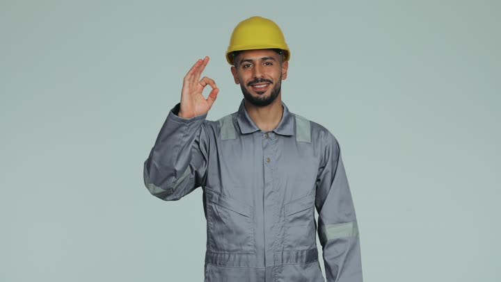 لباس رسمي للعمل في القطاع الهندسي، أعمال الإصلاح وورشات الصيانة في الإمارات العربية المتحدة, مهندس فني عربي خليجي اماراتي يرتدي الخوذة وبدلة السلامة يرفع يده للعبير عن الجودة ووالامتياز بإيماءات السعادة والسرور، المهن والوظائف الشبابية، خلفية بيضاء