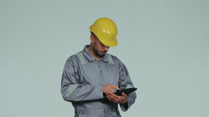 Integrating modern technologies into practical life, formal dress for work in the engineering sector, repair work and maintenance workshops in the UAE, an Arab Gulf Emirati technical engineer wearing a helmet and safety suit managing his work via a mobile tablet, youth professions and jobs, white background