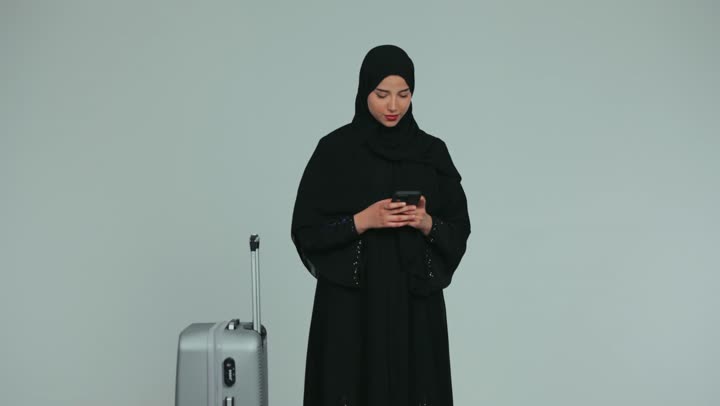 التقاط صورة سيلفي تذكارية بإيماءات السرور، الاستعداد لقضاء وقت ممتع, الاستمتاع بالرحلات السياحية في وقت العطلة, امرأة عربية خليجية إماراتية محجبة ترتدي العباءة تمسك بيدها حقيبة سفر رمادية تقوم باستخدام الهاتف المحمول, خلفية بيضاء