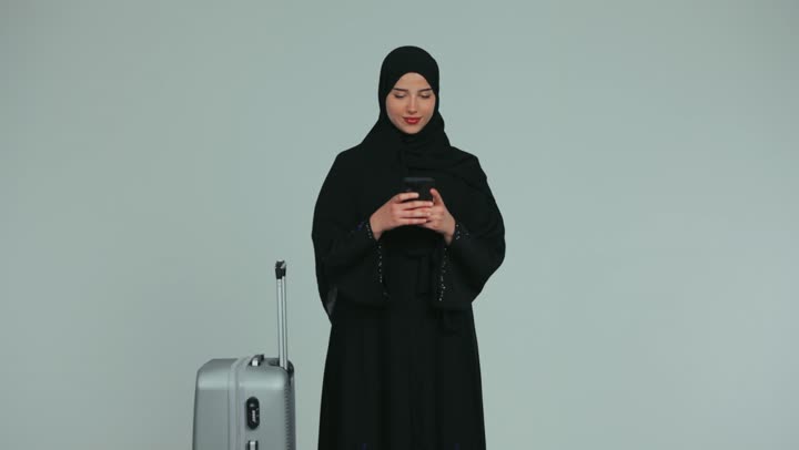 Tourism and hotel applications display, blank green chroma key screen, getting ready to have a good time, enjoying tourist trips during vacation time, veiled Emirati Gulf Arab woman wearing abaya holding gray suitcase using mobile phone, white background