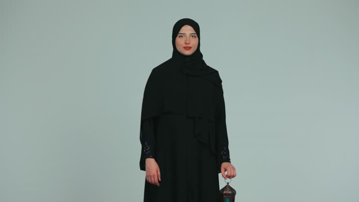 الإستعداد لقدوم شهر رمضان المبارك، أجواء السكينة والروحانية، الاحتفال بالمناسبات الدينية، شابة عربية خليجية اماراتية محجبة ترتدي العباءة تحمل بيدها فانوس معدني، النظر إلى الكاميرا بايماءات السعادة والسرور، خلفية بنفسجية