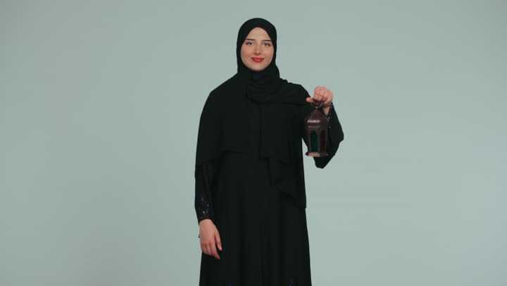 الإستعداد لقدوم شهر رمضان المبارك، أجواء السكينة والروحانية، الاحتفال بالمناسبات الدينية، شابة عربية خليجية اماراتية محجبة ترتدي العباءة تحمل بيدها فانوس معدني، النظر إلى الكاميرا بايماءات السعادة والسرور، خلفية بنفسجية