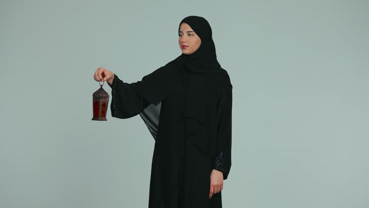 Preparing for the holy month of Ramadan, an atmosphere of serenity and spirituality, celebrating religious occasions, a young veiled Emirati Gulf Arab woman wearing an abaya holding a metal lantern and pointing at it with her index finger, white background