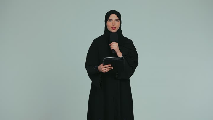 Presenting a lecture using a modern technical device, working in radio and news broadcasts, a veiled young Emirati Gulf Arab woman wearing an abaya holding a loudspeaker and a tablet, giving lectures and speeches and holding training workshops, women's professions and jobs in the Emirates, white background