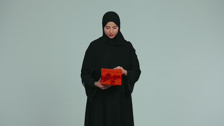 تقديم الهدايا بين الأهل والأصدقاء، التعبير عن المحبة والألفة، امرأة عربية خليجية اماراتية محجبة ترتدي عباءة تمسك بيدها علبة حمراء وتنظر إلى الكاميرا بإيماءات السعاده والسرور، خلفية بيضاء