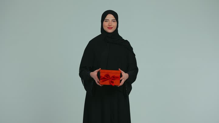 تقديم الهدايا بين الأهل والأصدقاء، التعبير عن المحبة والألفة، امرأة عربية خليجية اماراتية محجبة ترتدي عباءة تمسك بيدها علبة حمراء وتنظر إلى الكاميرا بإيماءات السعاده والسرور، خلفية بيضاء