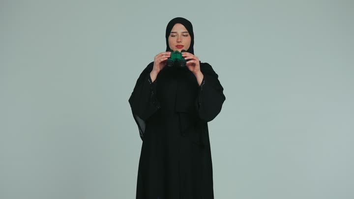 Observing and watching distant objects, future vision and setting goals and ambitions, a veiled Emirati Gulf Arab woman wearing an abaya holding binoculars in her hands and looking through them with gestures of pleasure, white background
