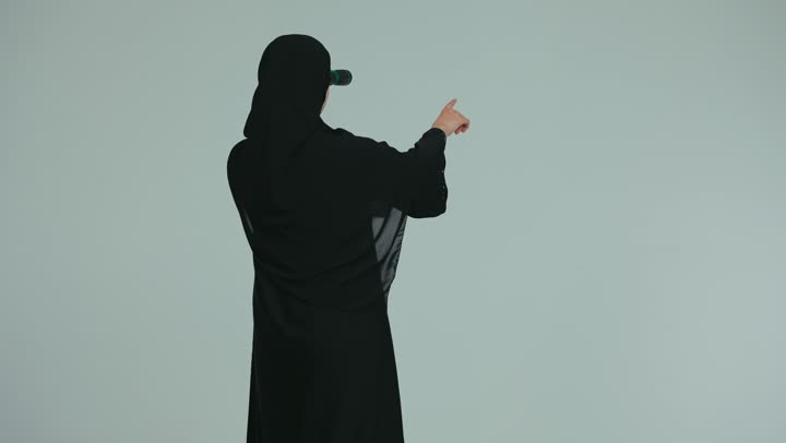 Pointing with the index finger at something, observing and watching distant things, future vision and setting goals and ambitions, a photograph from behind of a veiled young Arab Gulf Emirati woman wearing an abaya holding binoculars in her hands and looking through them, white background