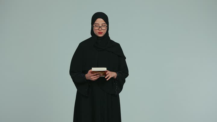 Spending free time learning and culture, interest in browsing useful books, love of reading, a veiled Emirati Gulf Arab woman wearing glasses and an abaya reading a book carefully and with concentration, culture of self-education, white background