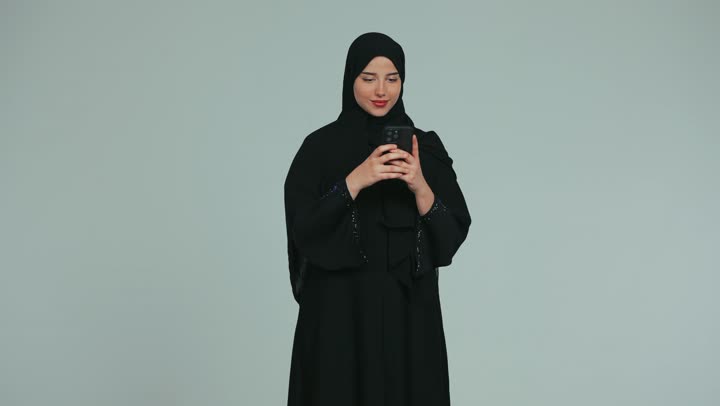 Taking souvenir selfies, taking advantage of modern technological devices in free time, a veiled Emirati Gulf Arab woman wearing an abaya using a mobile phone with gestures of happiness and pleasure, chatting and communicating with family and friends via mobile, white background