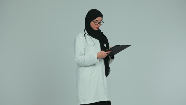 Women's professions and jobs, following up on patients' medical records, viewing test results, providing consultations in medical centers in the Emirates, working in the health sector, a veiled Emirati Gulf Arab female doctor wearing a medical coat and glasses taking notes, white background