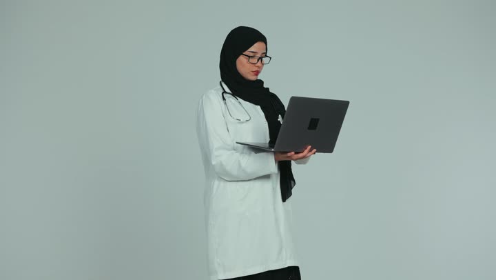 Conducting remote medical consultations, health professions and jobs, providing services to patients efficiently, using technology in research and development, an Arab Gulf Emirati female doctor wearing a white coat and glasses working on a laptop, white background