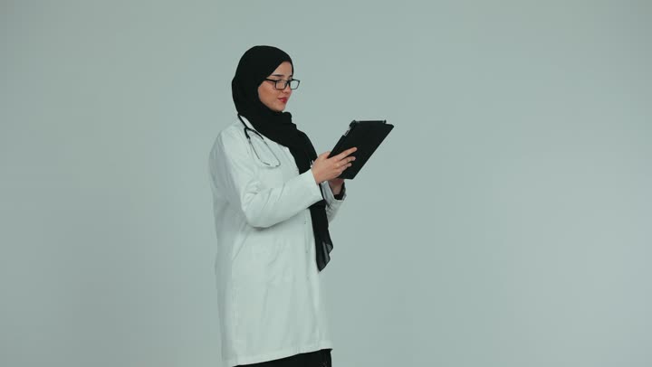 اجراء الاستشارات الطبيه عن بعد، مهن ووظائف صحية، تقديم الخدمات للمرضى بكل كفاءه، استخدام التكنولوجيا في البحث والتطوير، طبيبة عربية خليجية اماراتية ترتدي المعطف الابيض والنظارة تقوم بالعمل عبر جهاز التابلت، خلفيه بيضاء