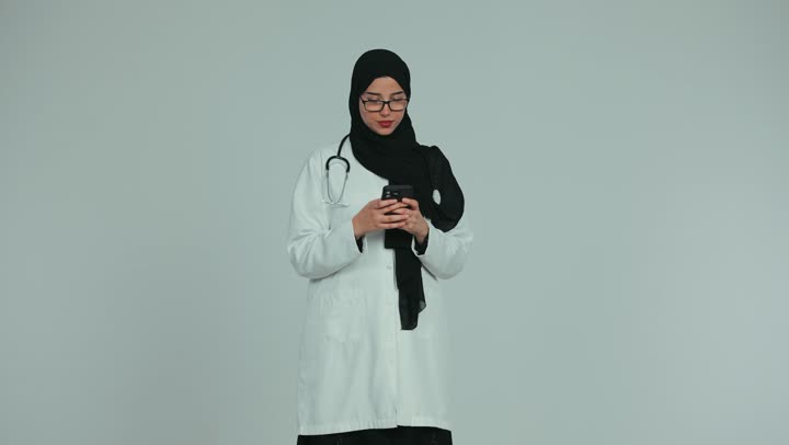 Health professions and jobs, providing services to patients efficiently, using technology in research and development, conducting remote medical consultations, an Arab Gulf Emirati female doctor wearing a white coat and glasses making a video call via mobile phone, white background