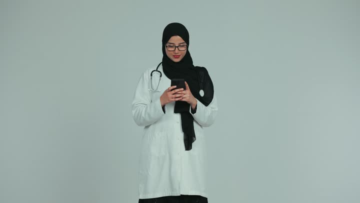 Providing services to patients efficiently, displaying a blank green chroma key screen, using technology in research and development, conducting remote medical consultations,  Arab Gulf Emirati female doctor wearing a white coat and glasses and working on a mobile phone, health professions and jobs, white background