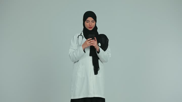 عرض شاشة كروما خضراء فارغة، تقديم الخدمات للمرضى بكل كفاءه، استخدام التكنولوجيا في البحث والتطوير، اجراء الاستشارات الطبيه عن بعد، لطبيبة عربية خليجية اماراتية ترتدي المعطف الابيض والسماعة الطبية وتعمل على الهاتف المحمول، مهن ووظائف صحية، خلفيه بيضاء