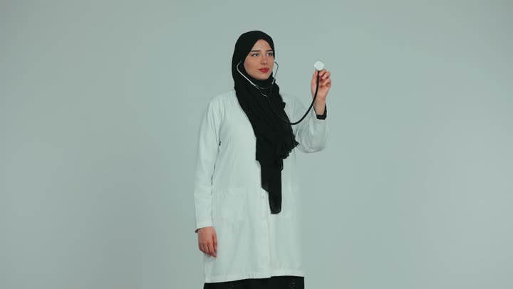 Working in the healthcare sector in the United Arab Emirates, a veiled female Emirati Gulf Arab doctor wearing a white medical coat performing a heartbeat examination with a stethoscope, health and healthcare concept, white background.