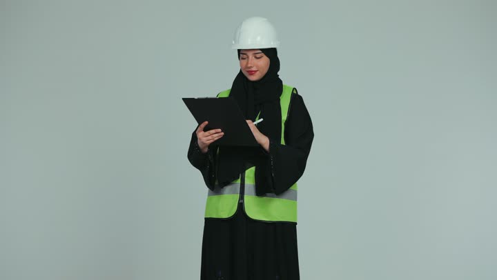Working in the engineering field, the concept of engineering and architectural construction, supervising and auditing work, a veiled Arab Gulf Emirati female engineer wearing a helmet and a protective vest taking notes on engineering plans and projects, white background.
