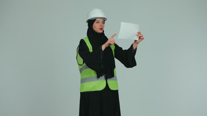 مفهوم الهندسة و الإنشاءات المعمارية, الإشراف على الأعمال وتدقيقها، مهندسة عربية خليجية اماراتية محجبة ترتدي الخوذة وسترة الحماية تقوم بتدقيق المخططات والمشاريع الهندسية، العمل في المجال الهندسي, خلفية بيضاء.