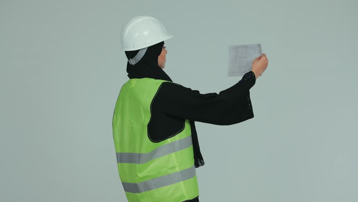Concept of engineering and architectural construction, supervision and inspection of works, veiled female Emirati Gulf Arab engineer wearing helmet and protective vest inspecting engineering plans and projects, working in the engineering field, white background.