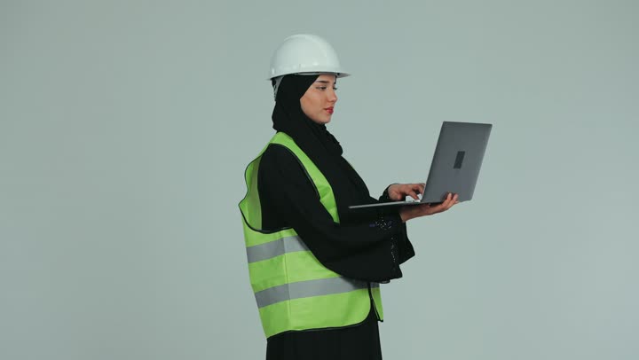 إدارة المشاريع باستخدام التقنيات الحديثة، بناء الوطن بسواعد أبنائه, مهندسة عربية خليجية اماراتية محجبة ترتدي الخوذة وسترة الحماية تقوم بمتابعة وتدقيق المخططات الإنشائية باستخدام الحاسوب المحمول، العمل في القطاع الهندسي، خلفية بيضاء