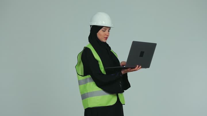إدارة المشاريع باستخدام التقنيات الحديثة، بناء الوطن بسواعد أبنائه, مهندسة عربية خليجية اماراتية محجبة ترتدي الخوذة وسترة الحماية تقوم بمتابعة وتدقيق المخططات الإنشائية باستخدام الحاسوب المحمول، العمل في القطاع الهندسي، خلفية بيضاء