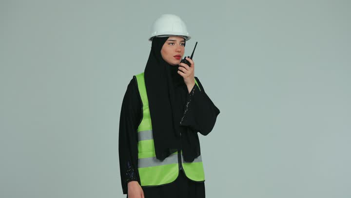 إعطاء أوامر خاصة بالعمل، مهندسة عربية خليجية اماراتية محجبة ترتدي الخوذة وسترة الحماية تقوم بالتحدث عبر الجهاز اللاسلكي  للإشراف على الأعمال ومتابعتها، المخططات والمشاريع الهندسية، مفهوم الهندسة و الإنشاءات, الغشارة بإصبع السبابة إلى شيء ما, خلفية بيضاء.