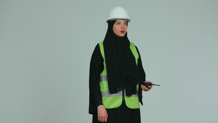 Giving work orders, a veiled Emirati Gulf Arab female engineer wearing a helmet and a protective vest talking on a walkie-talkie to supervise and follow up on work, engineering plans and projects, engineering and construction concept, pointing with index finger at something, white background.