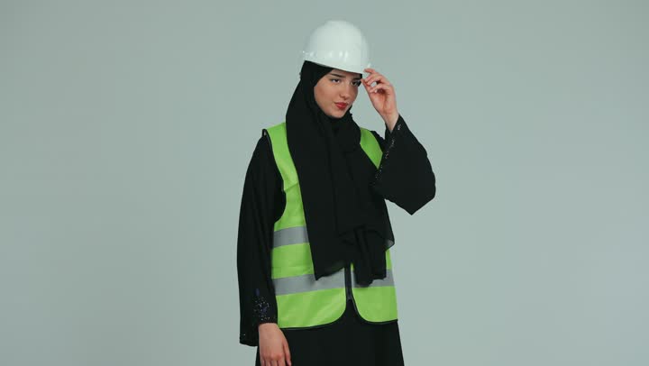 الوقوق بأيدي متقاطعة والنظر إلى الكاميرا، الإشراف على الأعمال وتدقيقها، مهندسة عربية خليجية اماراتية محجبة ترتدي الخوذة وسترة الحماية، المخططات والمشاريع الهندسية، مفهوم الهندسة و الإنشاءات, العمل في المجال الهندسي, خلفية بيضاء.