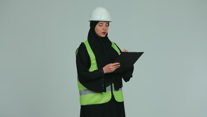 الإشراف على الأعمال وتدقيقها، مهندسة عربية خليجية اماراتية محجبة ترتدي الخوذة وسترة الحماية تحمل بيدها المخططات والمشاريع الهندسية، مفهوم الهندسة و الإنشاءات, العمل في المجال الهندسي, خلفية بيضاء.