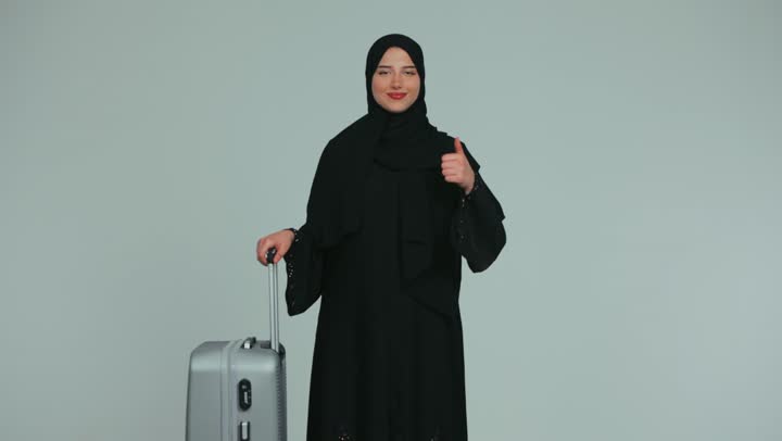 النظر إلى الكاميرا بإيماءات السرور، الاستعداد لقضاء وقت ممتع, الاستمتاع بالرحلات السياحية في وقت العطلة, امرأة عربية خليجية إماراتية محجبة ترتدي العباءة تمسك بيدها حقيبة سفر رمادية ترفع إصبع الإبهام بإيماءات الإعجاب والرضى, خلفية بيضاء