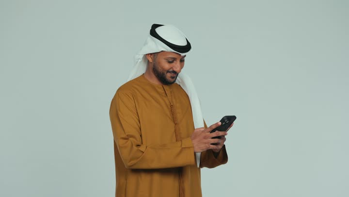 تصفح الانترنت وإنجاز المهام عن بعد، متابعه آخر أخبار السوشال ميديا، عقد اجتماع عمل، دمج التقنية والتكنولوجيا في الحياة اليومية, رجل عربي خليجي اماراتي يرتدي الكندورة والغترة يقوم باستخدام جهاز التابلت بتعابير السعادة والسرور, خلفية بيضاء