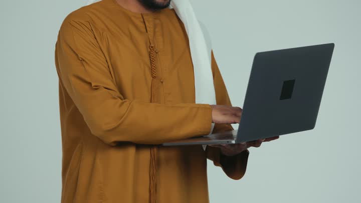 إدارة شؤون الأعمال وتنظيمها، استخدام التقنيات الحديثة المتطورة، عقد إجتماع عمل عبر الإنترنت، تصوير مقرب لرجل عربي خليجي سعودي يرتدي الكندورة والغترة يقوم بالعمل على الحاسب المحمول, دمج التكنولوجيا في الحياة العملية، خلفية بيضاء