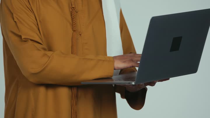 إدارة شؤون الأعمال وتنظيمها، استخدام التقنيات الحديثة المتطورة، عقد إجتماع عمل عبر الإنترنت، تصوير مقرب لرجل عربي خليجي سعودي يرتدي الكندورة والغترة يقوم بالعمل على الحاسب المحمول, دمج التكنولوجيا في الحياة العملية، خلفية بيضاء