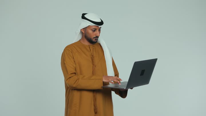 Greeting and saluting, managing and completing tasks using a modern technical device, close-up of an Emirati Gulf Arab man wearing a white kandura and ghutra conducting a business meeting via laptop, Emirati office jobs and professions, making a video call with family and friends, white background
