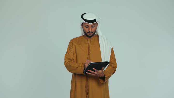 Browsing social media applications, using modern and advanced technology, using a tablet to complete work tasks, an Emirati Gulf Arab man wearing an Emirati kandura and ghutra holding a pen in his hand, writing on a tablet with gestures of seriousness and focus, white background