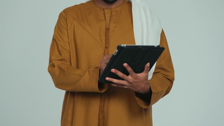 استعمال التكنولوجيا الحديثة، استخدام الجهاز اللوحي في إنجاز مهام العمل، تصفح تطبيقات التواصل الاجتماعي، تصوير مقرب لرجل عربي خليجي اماراتي يرتدي الكندورة والغترة الاماراتية يمسك بيده القلم يقوم بالكتابة على جهاز تابلت، خلفية بيضاء
