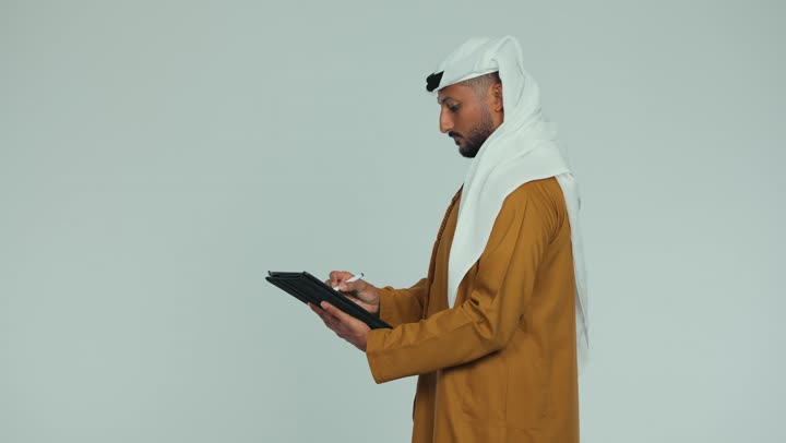 Using a tablet to complete work tasks, browsing social media applications, using modern technology, a side view of an Emirati Gulf Arab man wearing an Emirati kandura and ghutra, holding a pen in his hand, writing on a tablet with gestures of seriousness and concentration, white background