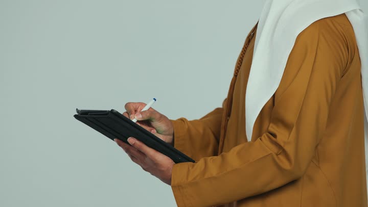 Using modern technology, using a tablet to complete work tasks, browsing social media applications, close-up side view of an Emirati Gulf Arab man wearing an Emirati kandura and ghutra holding a pen in his hand writing on a tablet, white background