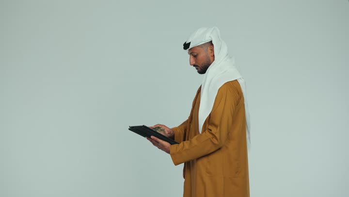 Watching favorite movies and series online, using modern technology, using a tablet to complete work tasks, side view of an Emirati Gulf Arab man wearing an Emirati kandura and ghutra holding a tablet in his hand browsing social media, white background