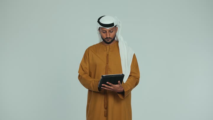 تصفح تطبيقات التواصل الاجتماعي، استعمال التكنولوجيا الحديثة والمتطورة، استخدام الجهاز اللوحي في إنجاز مهام العمل، رجل عربي خليجي اماراتي يرتدي الكندورة والغترة الاماراتية يمسك بيده القلم يقوم بالكتابة على جهاز تابلت بإيماءات الجدية والتركيز، خلفية بيضاء