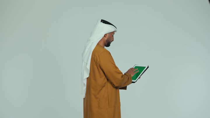 عرض شاشة كروما خضراء فارغة على الأجهزة التقنية، متابعة الأفلام والمسلسلات المفضلة عبر الإنترنت، استخدام الجهاز اللوحي في إنجاز العمل، تصوير من الجانب لرجل عربي خليجي اماراتي يرتدي الكندورة والغترة يمسك بيده التابلت يقوم بتصفح وسائل التواصل الإجتماعي، خلفية بيضاء