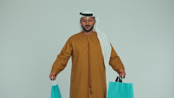 متعة التسوق في الإمارات، النظر إلى الكاميرا بإيماءات السعادة والسرور، الاستفادة من الحسومات والتصفيات، التجارية والهدايا, التبضع وأخذ الاحتياجات اللازمة، مواسم العروض والتخفيضات, رجل عربي خليجي اماراتي يرتدي الكندورة والغترة يمسك بيده أكياس ورقية ملونة، خلفية بيضاء