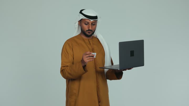Ease of purchasing and paying by bank credit card, the ability to pay bills with electronic cards, the use of modern devices and technologies, an Arab Gulf Emirati man wearing a kandura and a ghutra holding an ATM card in his hand and working on a laptop, white background