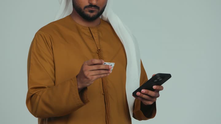 إمكانية تسديد الفواتير بالبطاقات الإلكترونية, استخدام الأجهزة والتقنيات الحديثة, سهولة الشراء والدفع عن طريق بطاقة الإئتمان البنكية، تصوير مقرب لرجل عربي خليجي اماراتي يرتدي الكندورة والغترة يمسك بيده بطاقة الصراف ويعمل على الهاتف المحمول, خلفية بيضاء