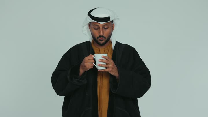 ارتداء الملابس الدافئة في فصل الشتاء، التلذذ والاستمتاع بتناول المشروب الساخن في الجو الشتوي البارد، الشعور بالصقيع والقشعريرة والتجمد، إيماءات تدل على البرد الشديد، رجل عربي خليجي اماراتي يرتدي الفروة و والغترة يقوم بشرب كوب من القهوة، خلفية بيضاء
